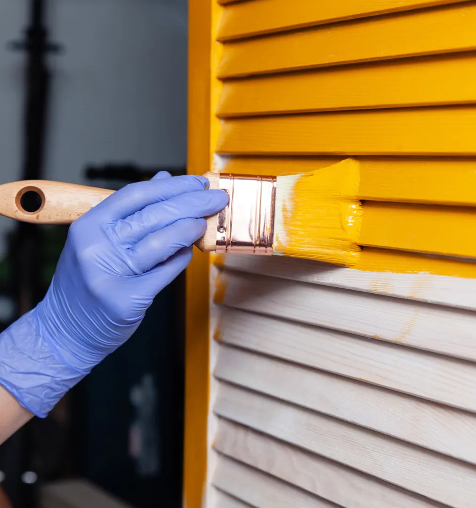 Gloved hand brushing yellow paint onto wooden shutters